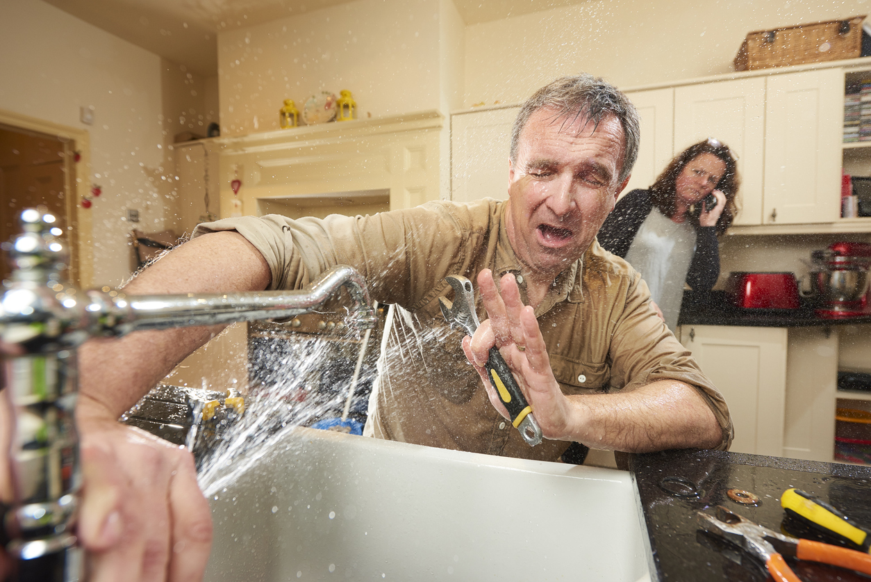 Man trying to stop a burst pipe under the sink while his wife calls a restoration company